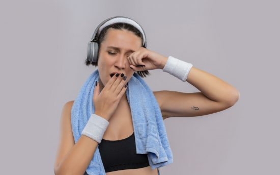 young fitness girl in sportswear with headphones on head and towel on her neck yawning covering mouth with hand wants to sleep standing over white background